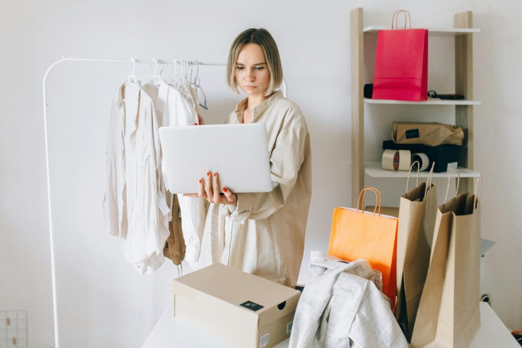 Woman using laptop for online shopping with clothes and bags around her, indoors.